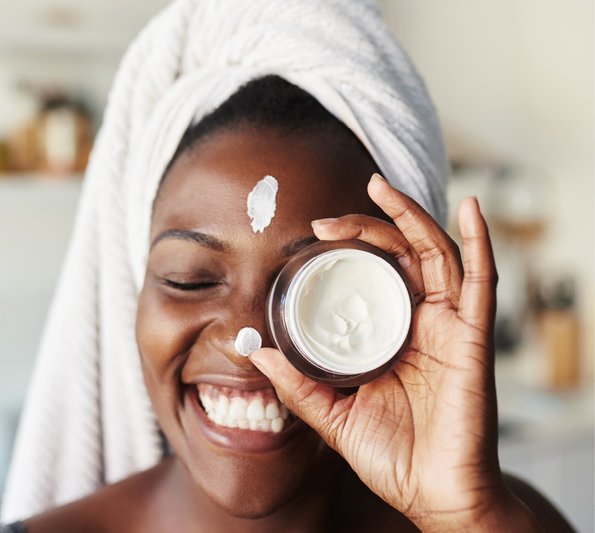 Cosmetic Services - young woman with dark skin, having her hair in a towel, she is holding a lotion in her hands in front of her left eye