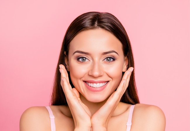 Rahn cosmetic news - young woman with a smile holding her hands next to her cheeks, the background is pink