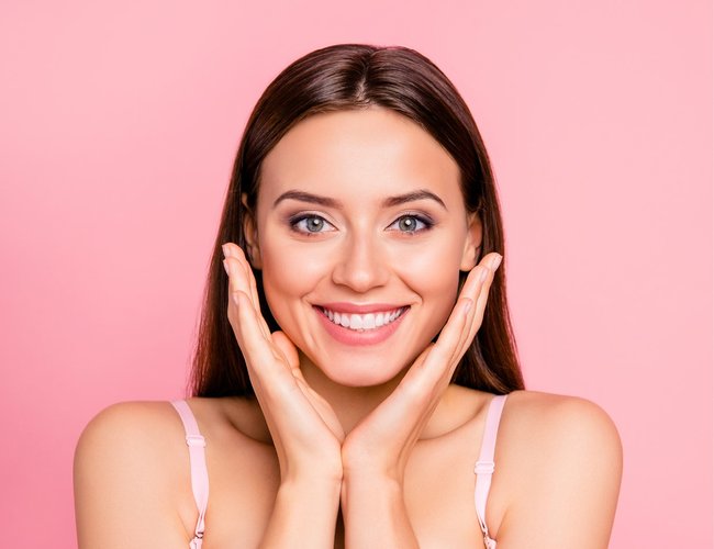 Rahn cosmetic news - young woman with a smile holding her hands next to her cheeks, the background is pink